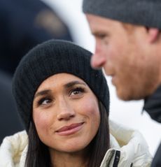 Meghan Markle wearing a black hat and white winter coat smiling up at Prince Harry, dressed in a black coat and hat