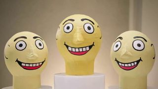 Three oversized, yellow papier-mâché heads with wide, staring eyes and broad, toothy grins are displayed on white pedestals against a neutral background.