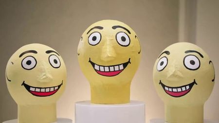 Three oversized, yellow papier-mâché heads with wide, staring eyes and broad, toothy grins are displayed on white pedestals against a neutral background.