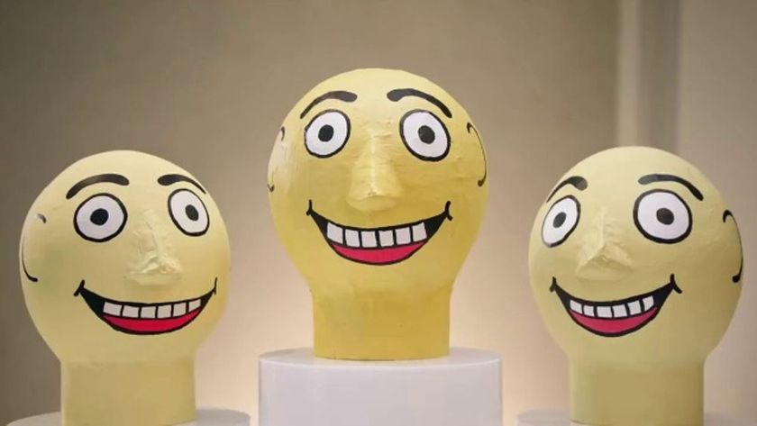 Three oversized, yellow papier-m&acirc;ch&eacute; heads with wide, staring eyes and broad, toothy grins are displayed on white pedestals against a neutral background.