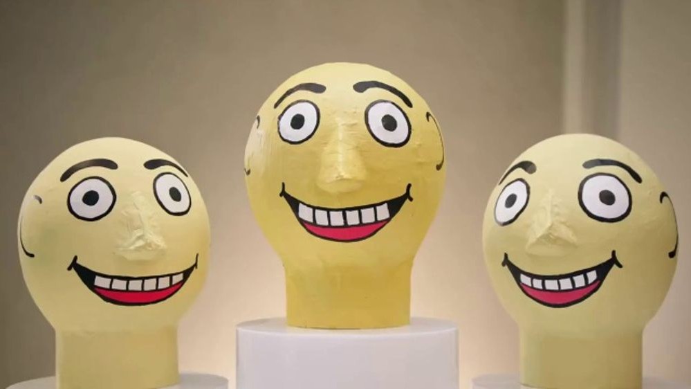 Three oversized, yellow papier-m&acirc;ch&eacute; heads with wide, staring eyes and broad, toothy grins are displayed on white pedestals against a neutral background.