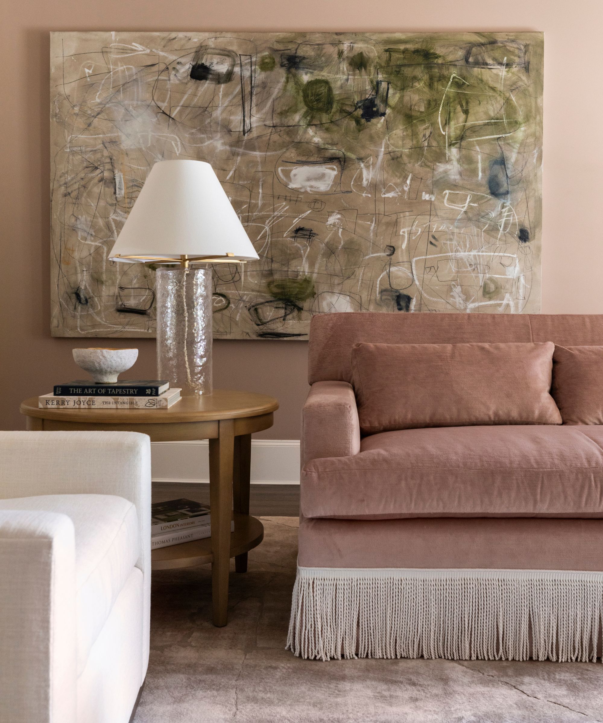 blush pink living room with a dusty pink velvet fringe sofa, white armchair, a large sideboard with artwork, and an oversized abstract painting on the back wall