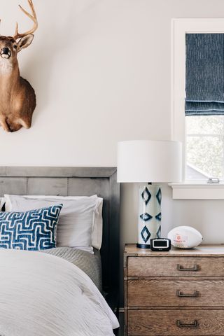 grey wooden bedhead with blue geometric cushion and lamp base and wooden chest of drawers and wall mounted deer head above the bed