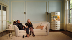 a large living room with two women sitting on a cream couch, a large bar cabinet to the left, and a side table to the right with a vase of flowers on it 