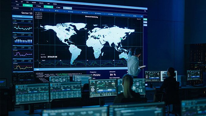 A control room with a huge display of maps being controlled at displays by several men and women.