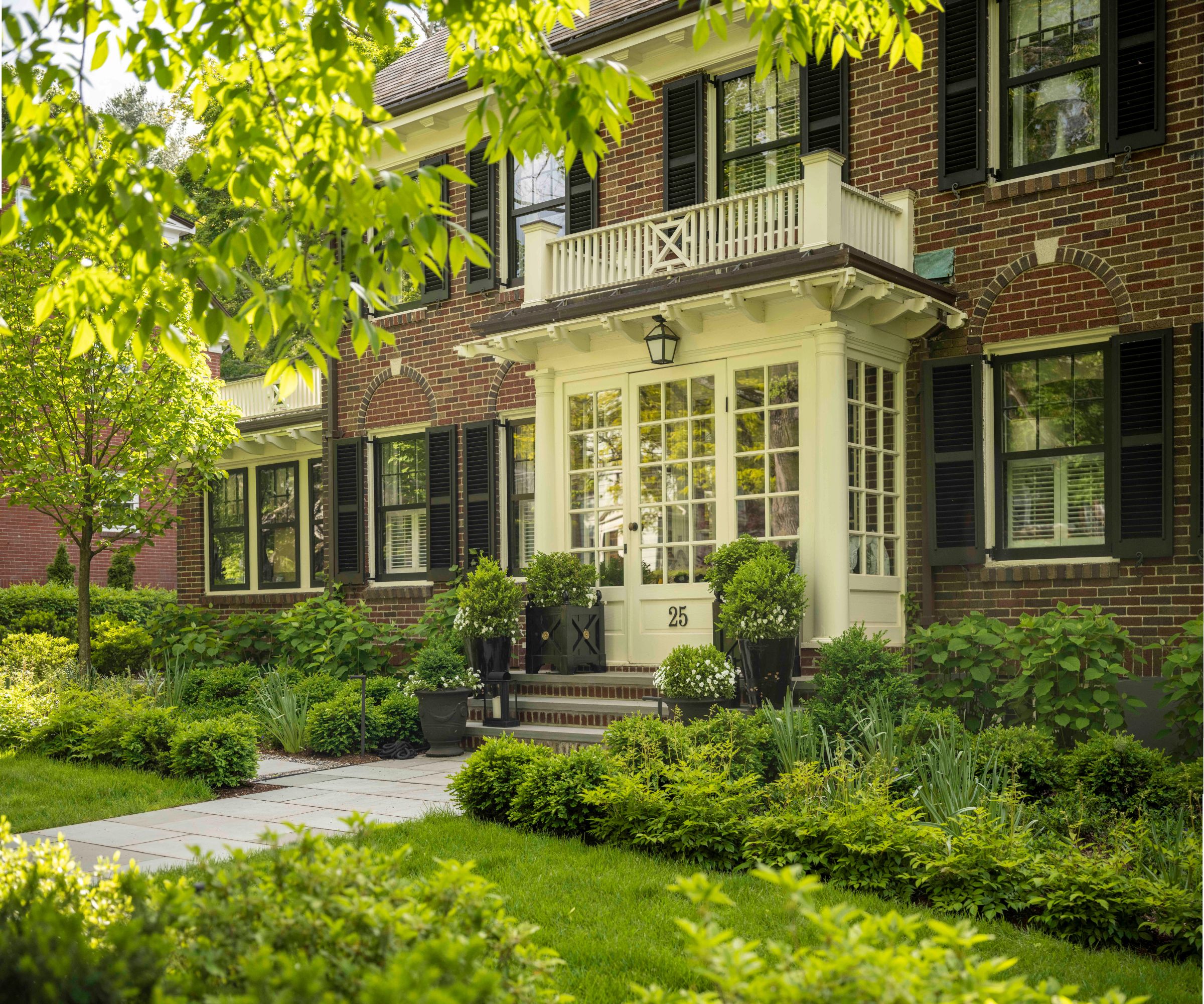 Grand red brick home with white balcony and front door and luscious greenery and potted plants
