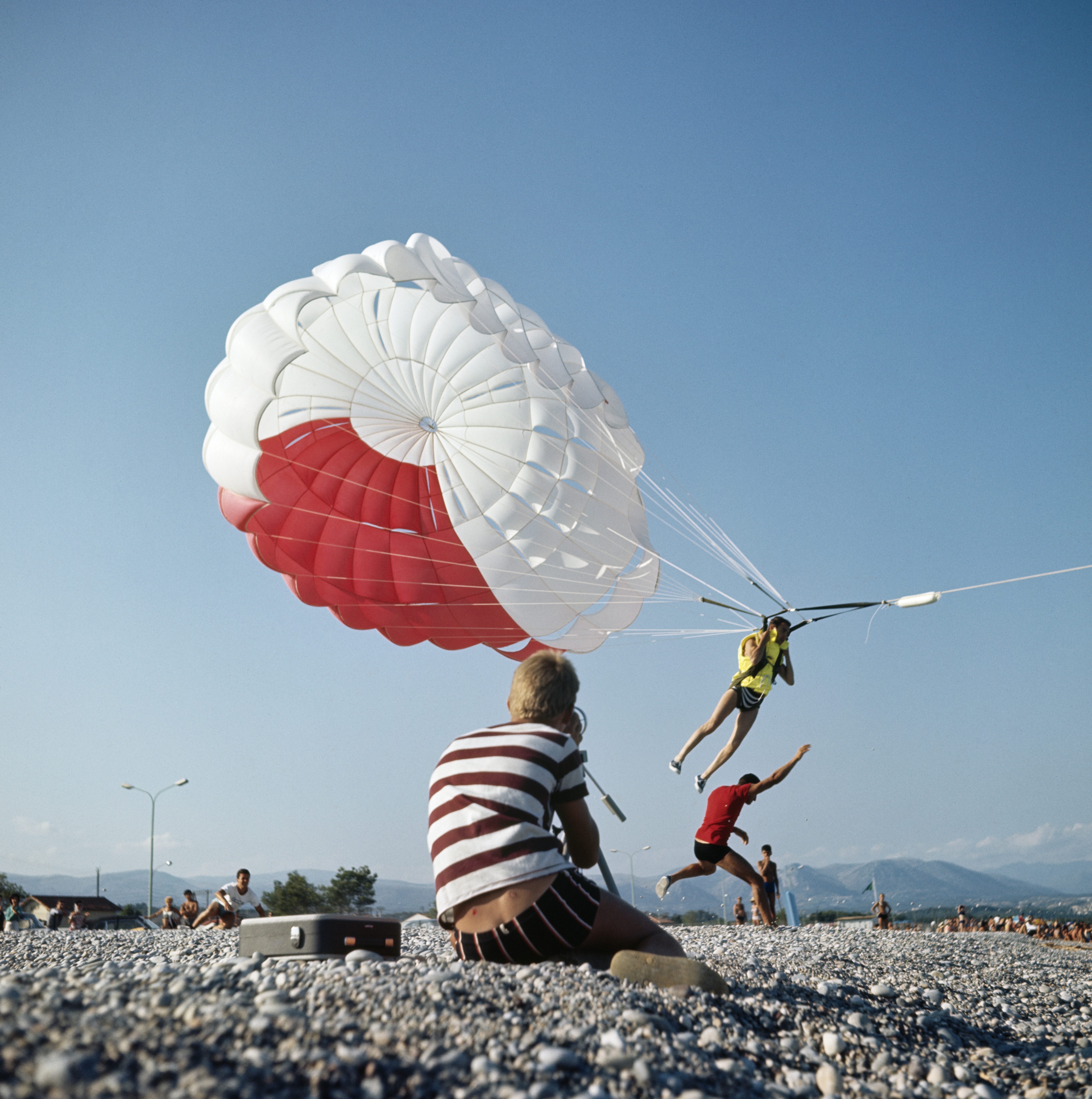A man in a striped top photographs a parasailer in a red and white canopy lifting off a pebble beach under a clear blue sky.