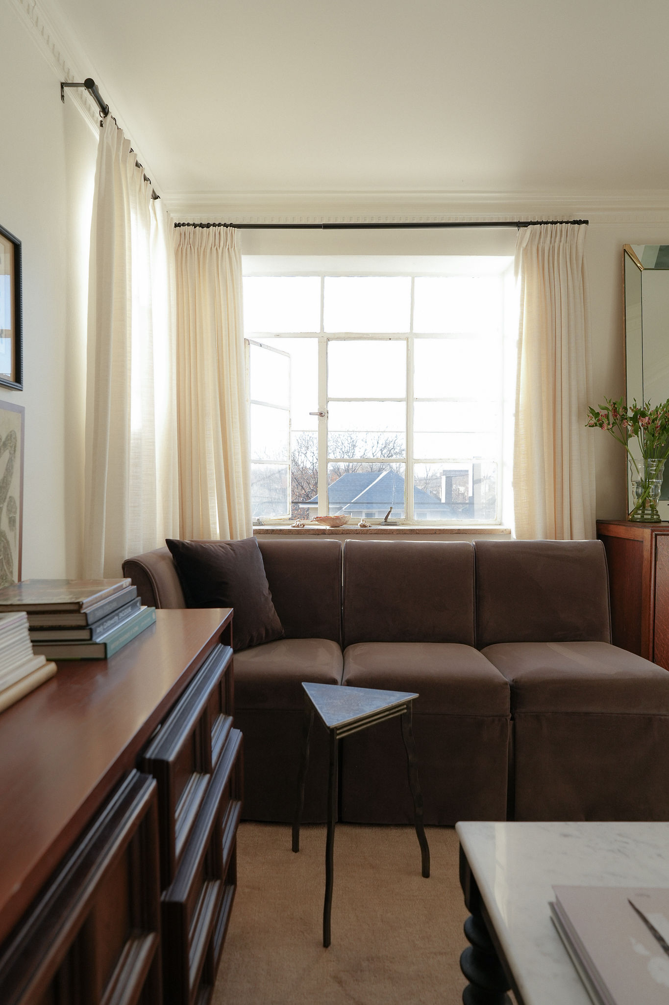 A white living room with white curtains on the window and a light brown rug. There is a dark brown sofa with a small accent table in front of it and a wooden console table in view.
