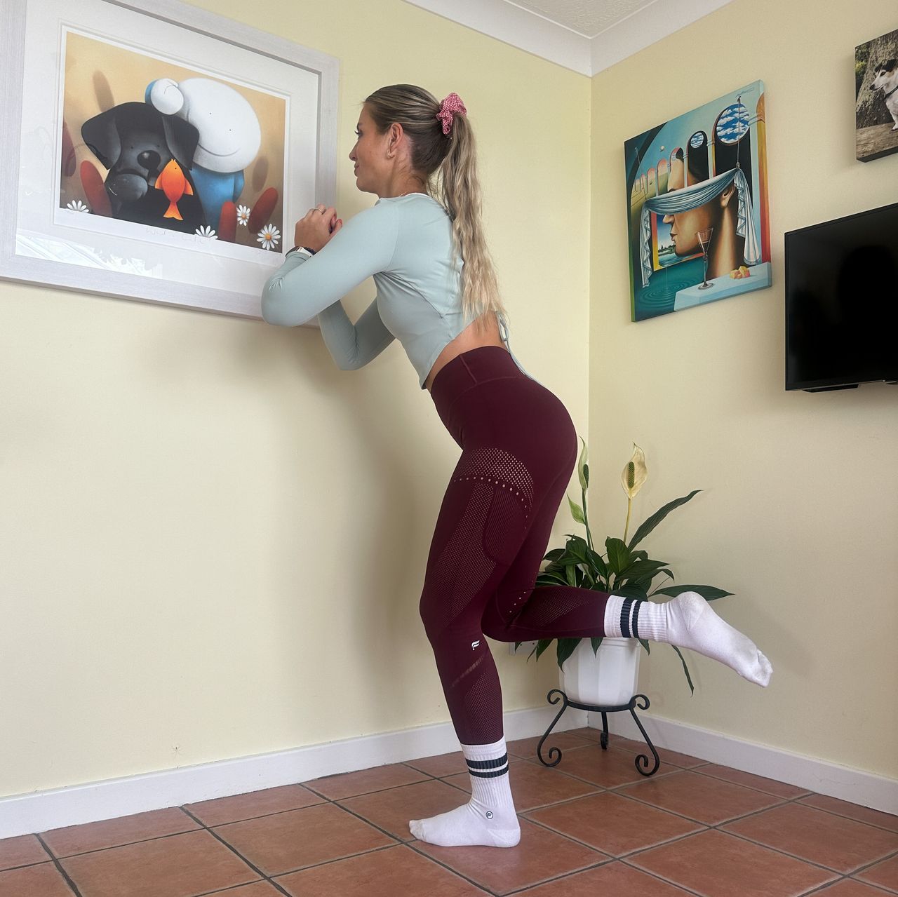 NASM qualified trainer Chloe Thomas performs a counterbalance squat in a room with cream colored walls and pictures on the walls. She is standing on one leg, leaning forward slightly and squatting down.