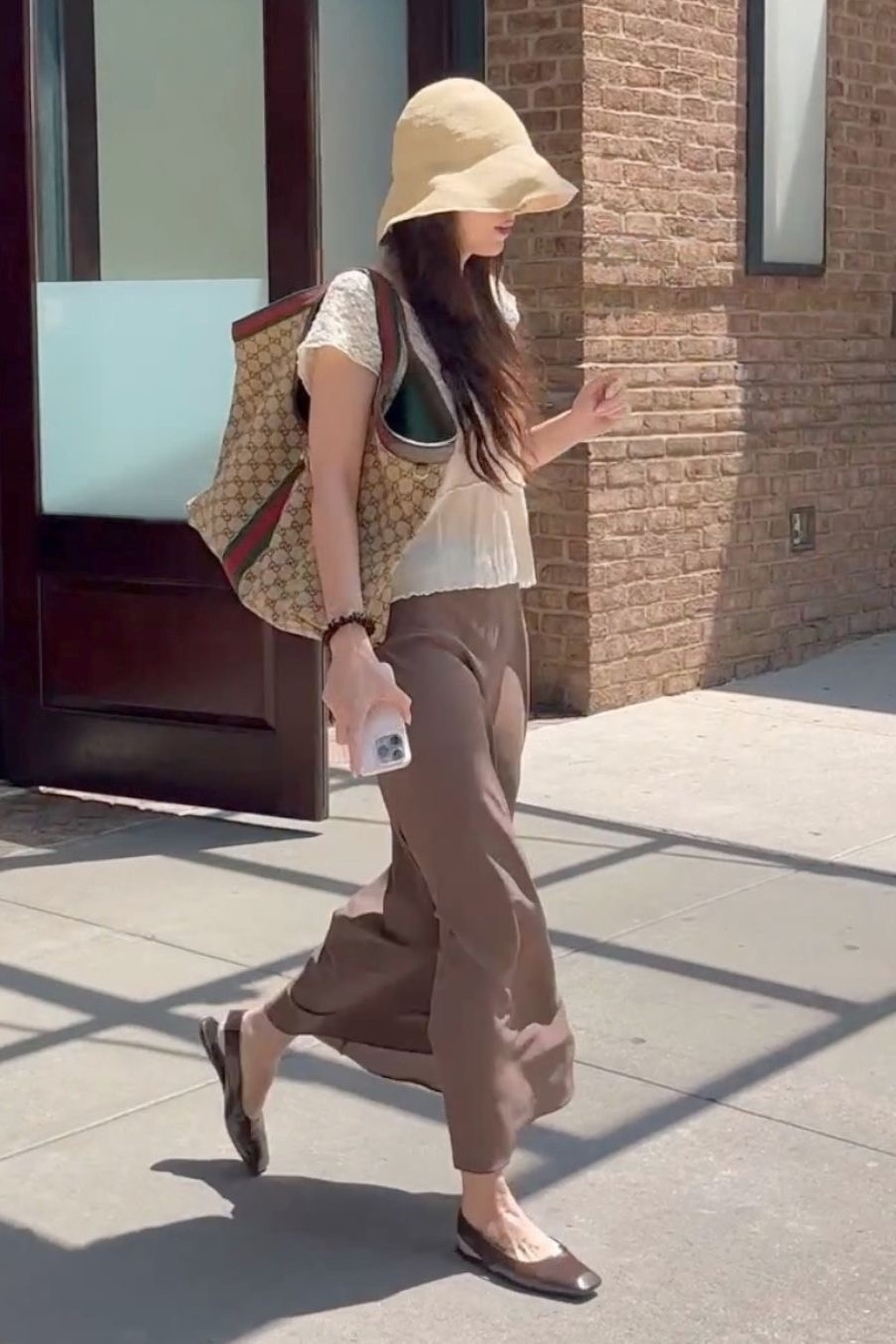Dakota Johnson steps outside wearing dark brown slingback flats with brown wide-leg trousers and white lace t-shirt. She completed her look with a Gucci bag and straw bucket hat.