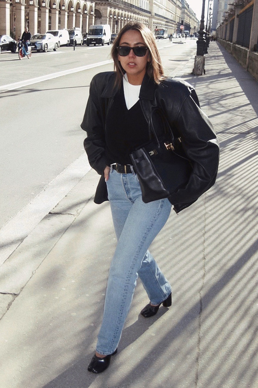 How to wear skinny jeans is shown in a street style photo of a woman walking down the sidewalk wearing black sunglasses, a black leather bomber jacket layered over a black v-neck sweater and white t-shirt styled with a black belt, light wash skinny jeans, black tabi pumps, and a black hermes bag