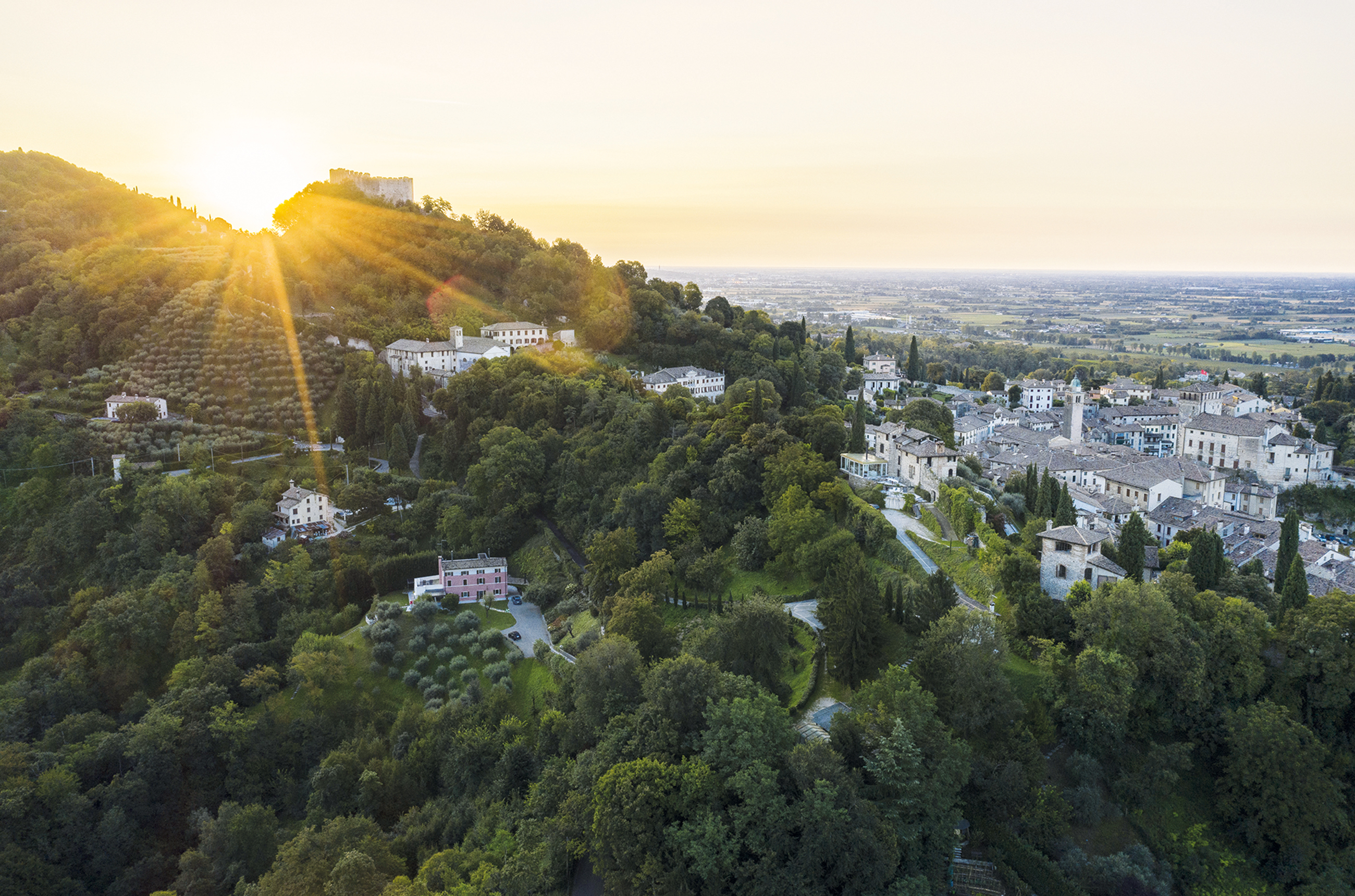 Birds-eye view shot of hills covered in trees with a small village of white and grey buildings to the right, at sunset