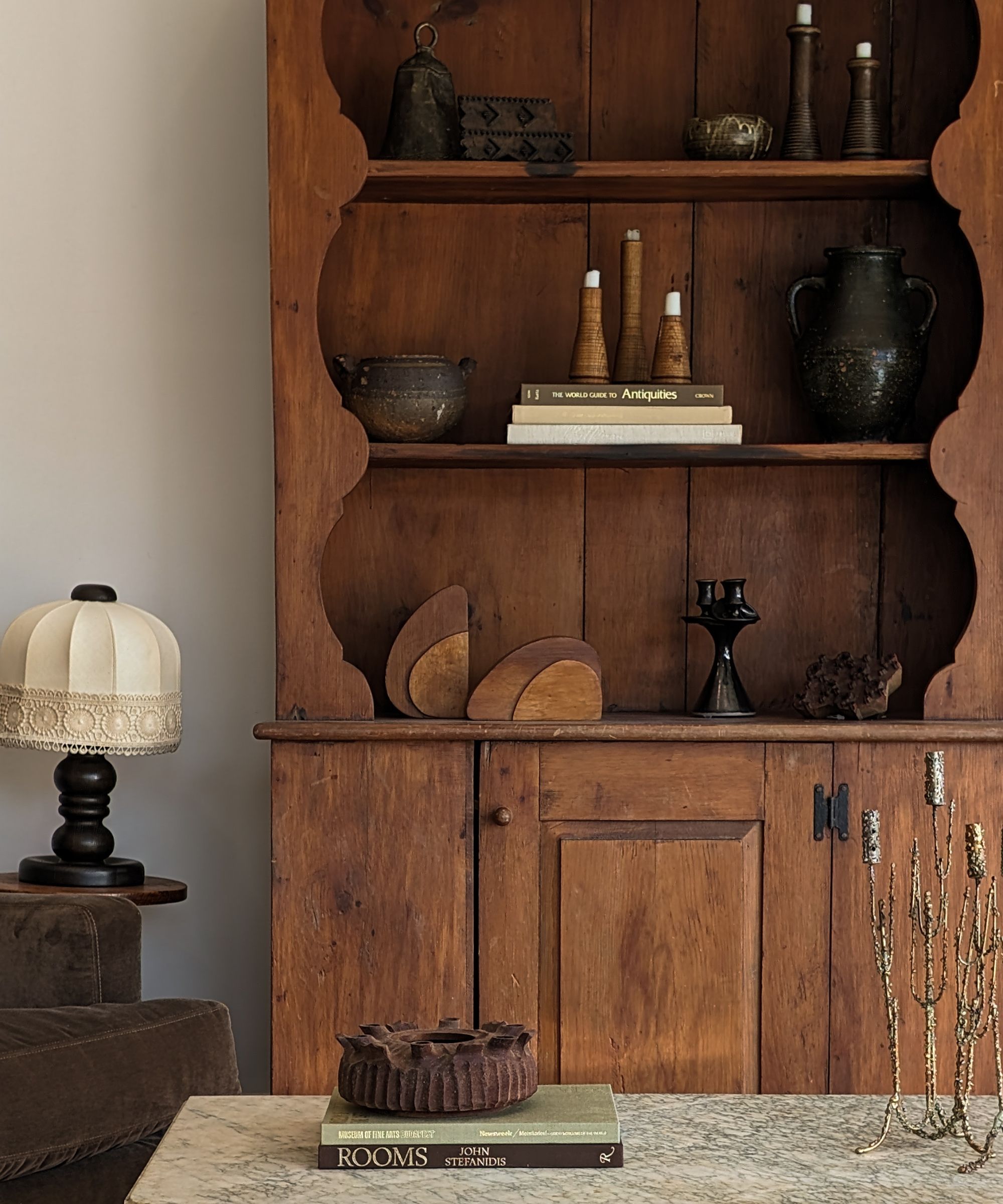 Antique wooden cabinet with scalloped detailing and open shelving, styled with pottery, candlesticks and stacked books, beside a small table lamp and marble-topped coffee table.