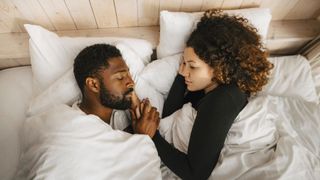 Man and a woman in bed together, under white bedsheets, holding hands