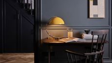 A yellow home office desk light with a large half circle shade and a curved gold stand on a wooden office desk against a dark navy wall
