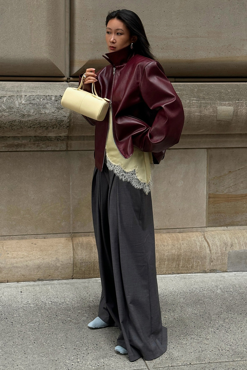 The best statement coats are shown in a street style photo of a woman standing on the sidewalk near a concrete building, wearing a burgundy red bomber jacket over a yellow camisole top styled with wide-leg gray trousers, blue heels, and a butter yellow barrel bag