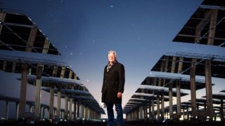 An older man with white hair and glasses wearing a dark jacket stands between large rows of solar panels under a night sky full of stars.