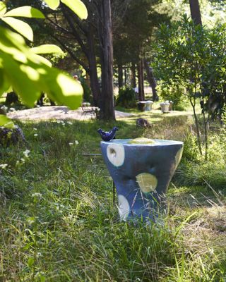 A colourful, freestanding bird bath in blue ceramic with white and yellow polka dots and a blue bird on top. Designed by Hung-Chung Lee and photographed in a garden surrounded by greenery
