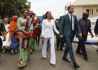Meghan Markle and Prince Harry walk in Nigeria with a delegation while Meghan wears a white flared suit