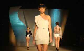 Model on runway wearing a cream, cold-shoulder dress, gathered at the thigh