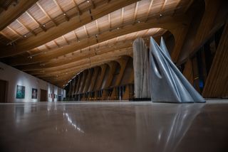 Interior of the building in a large space showcasing the curved wood roof details, wall art (on the left wall) and tall grey sculptured on the right