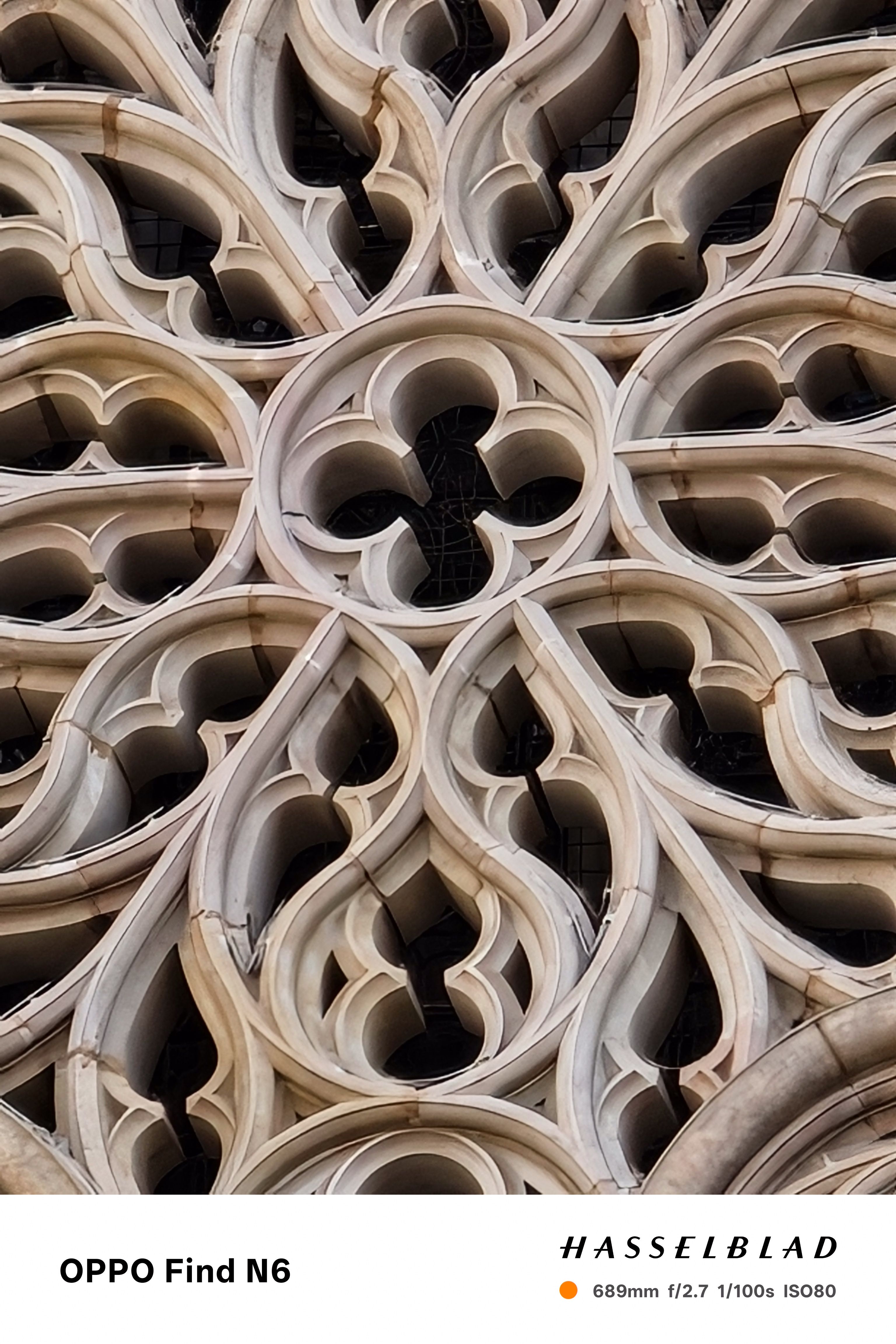 An extreme close-up of the intricate stone tracery of a Gothic rose window. The shot highlights the repetitive, flowing "flamboyant" patterns and the weathered texture of the light-colored sandstone.