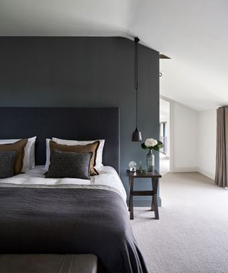 Large bedroom with graphite painted wall behind bed, dark gray headboard and blanket, white bedding, brown and gray cushions, cream carpet, dark wooden bedside table, black hanging pendant over table, en-suite bathroom to the right of bed