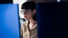 Female cyber security worker with glasses sitting at a workstation in an office environment.