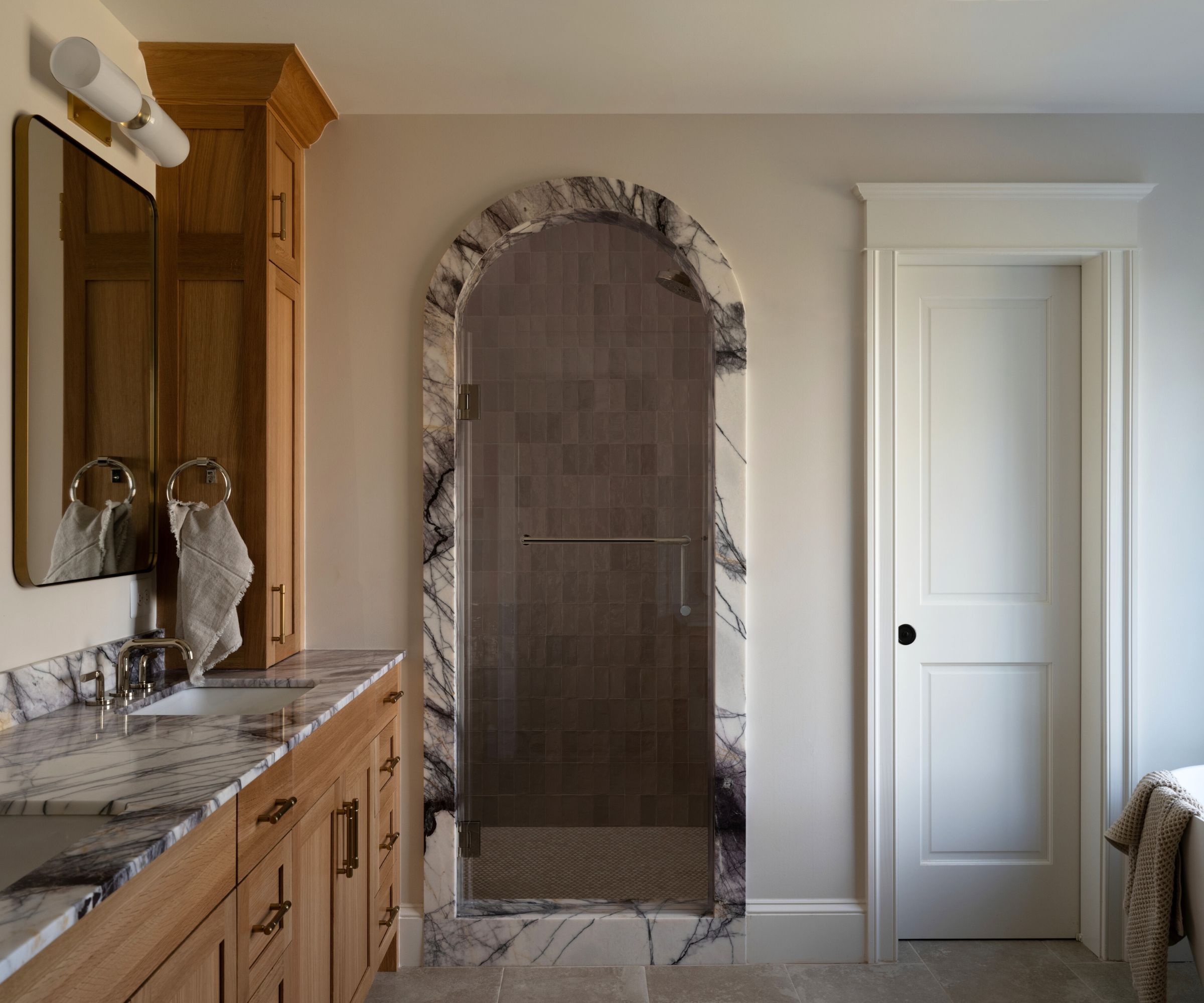 A warm neutral bathroom with a wooden vanity and marble edging surrounding the shower nook archway