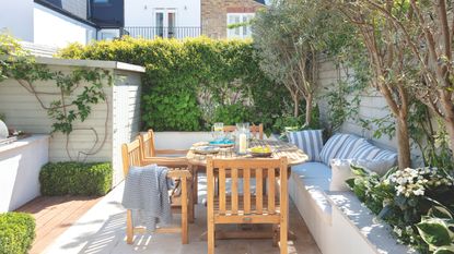 Wooden garden furniture on patio area in garden surrounded by white raised planters and seating and wall-climbing plants