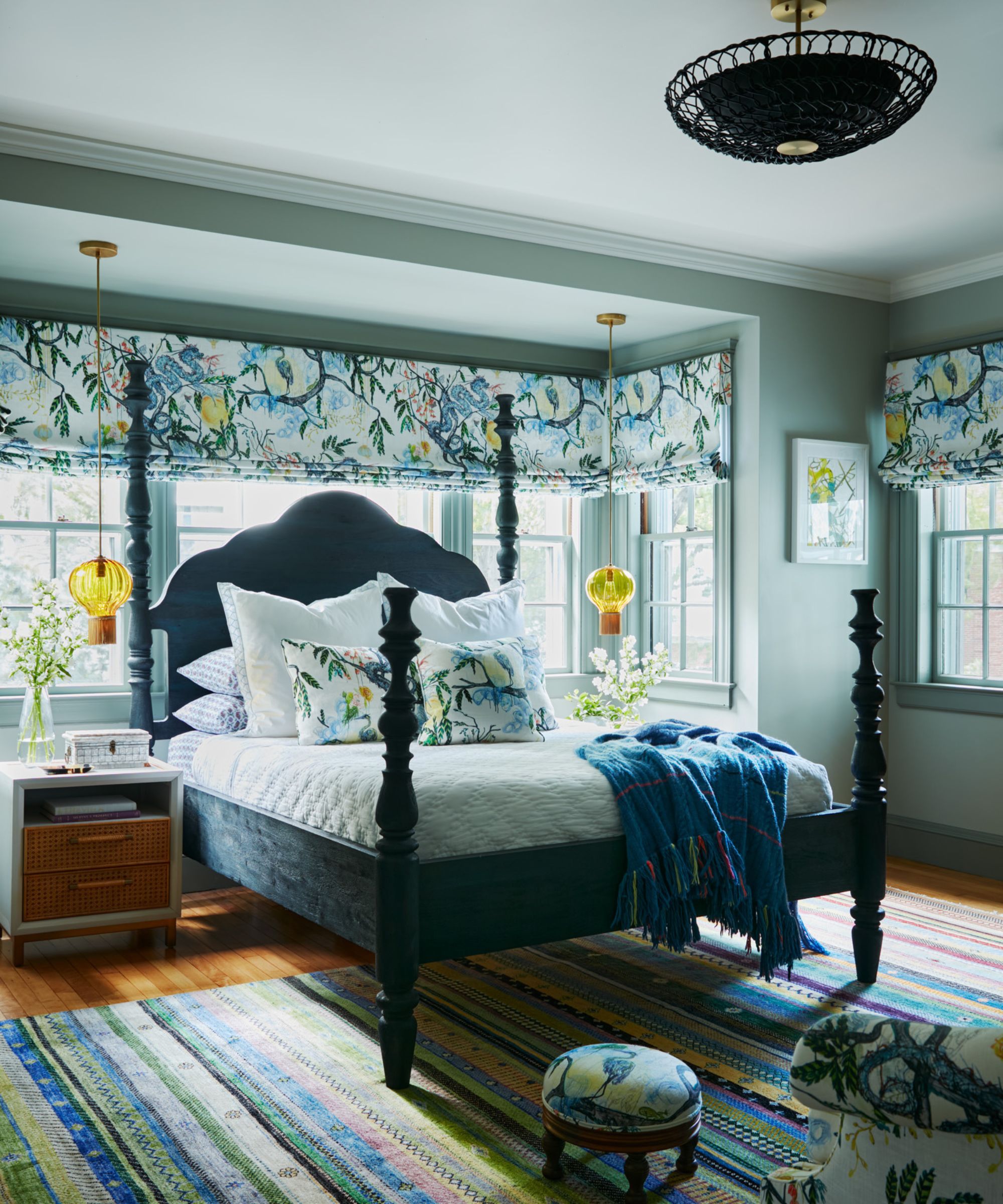A bedroom with gray-green walls and a lighter ceiling, large windows with blue floral roman blinds, a four-poster bed, and a striped rug.