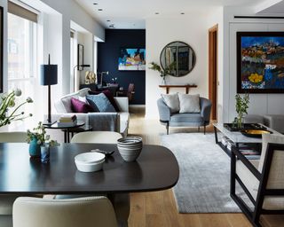 Open-plan living and family room space, dark navy feature wall, dark wooden flooring, zones area for seating, dining and working, gray sofa and lounge chair, gray rug and white marble coffee table, colorful artwork on walls, rounded mirror, black wood dining table