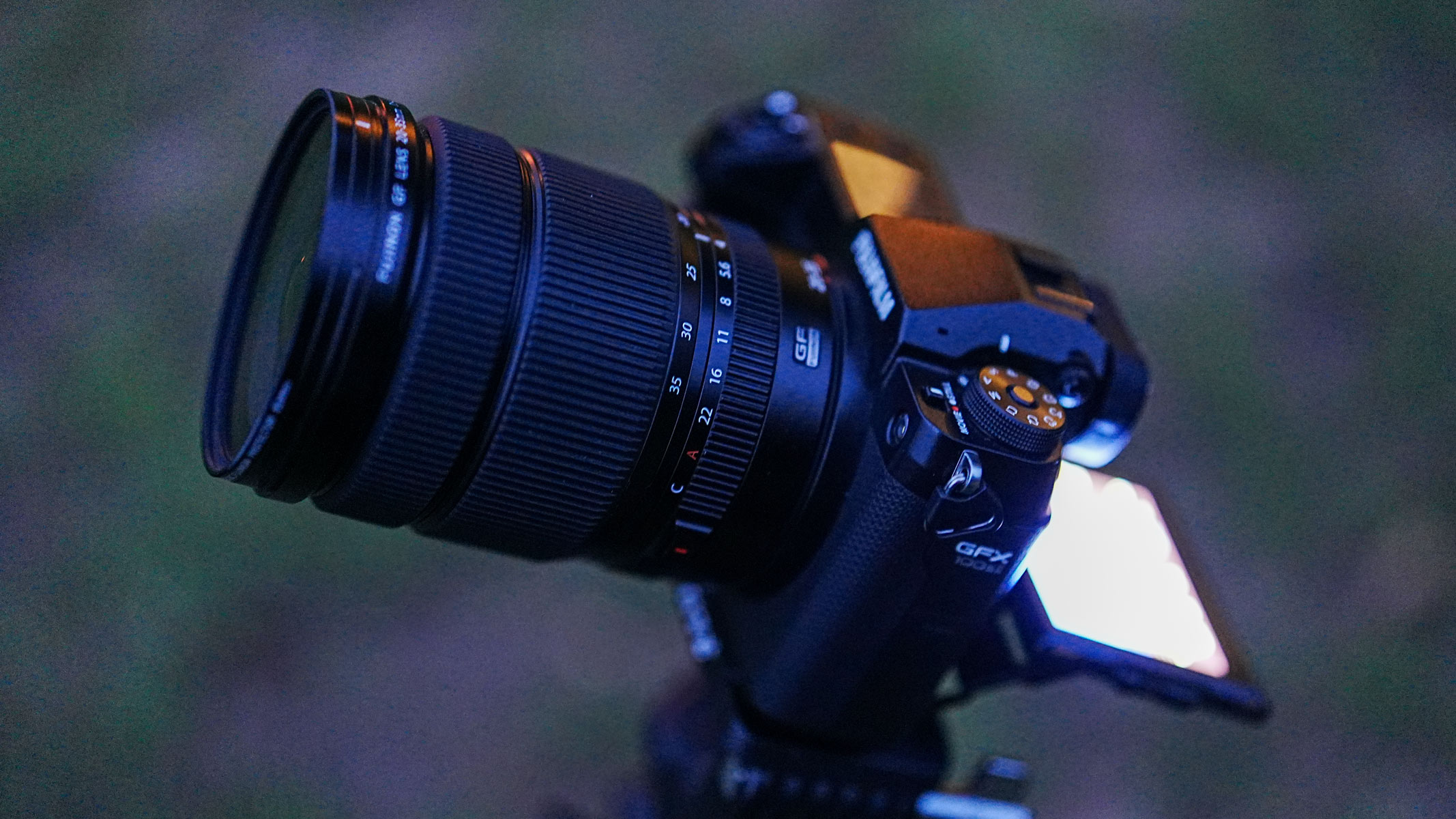 The Fujifilm GFX100S II with the Fujifilm GF 20-35mm f/4 R WR lens attached with the LCD screen extended and glowing, with grass in the background.