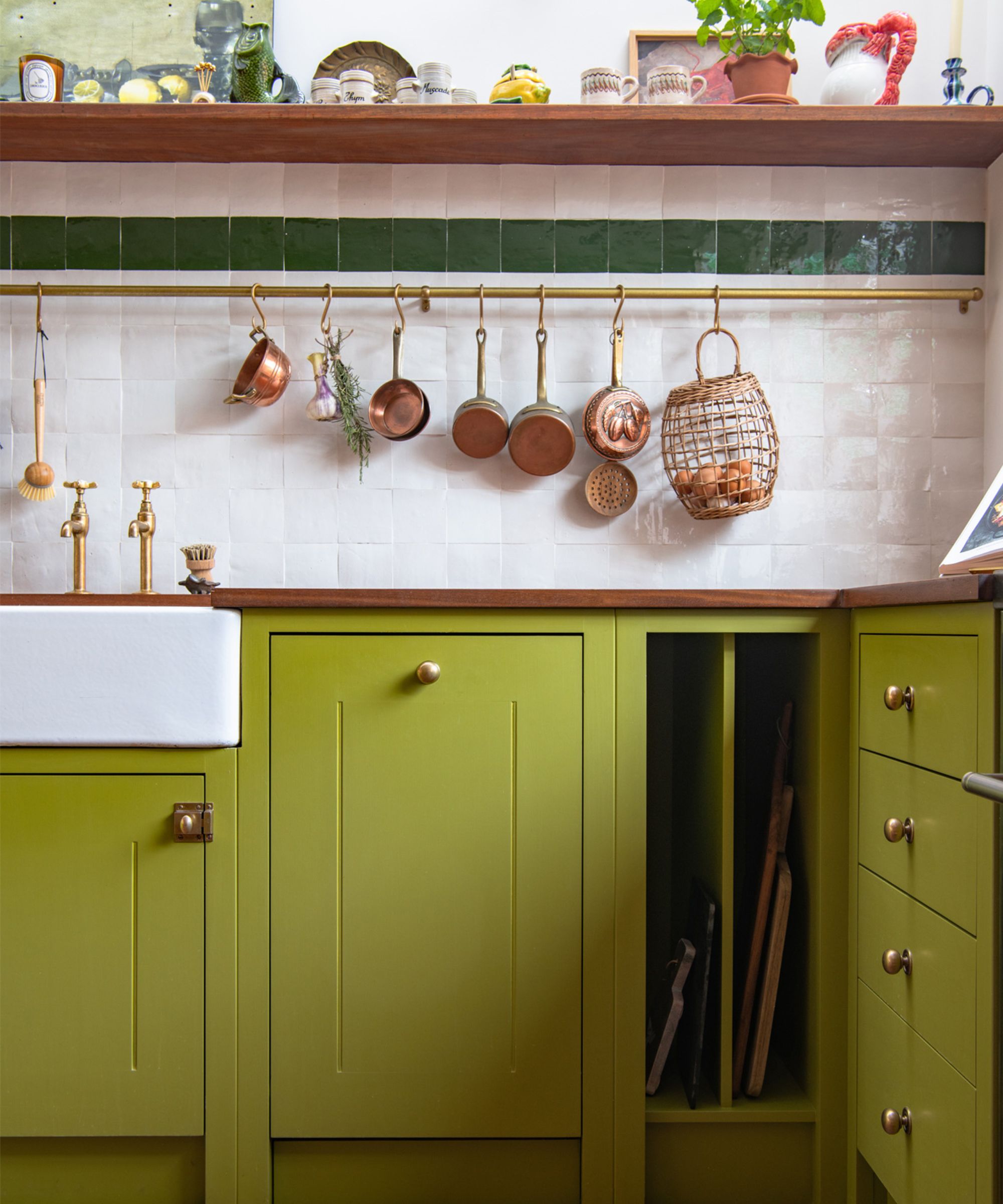 Kitchen with citron green cabinetry and gold handles, a ceramic white sink, tiled backsplash in cream with a green stripe, a pot rail with copper pans hanging from it, and a shelf of vintage glasses.