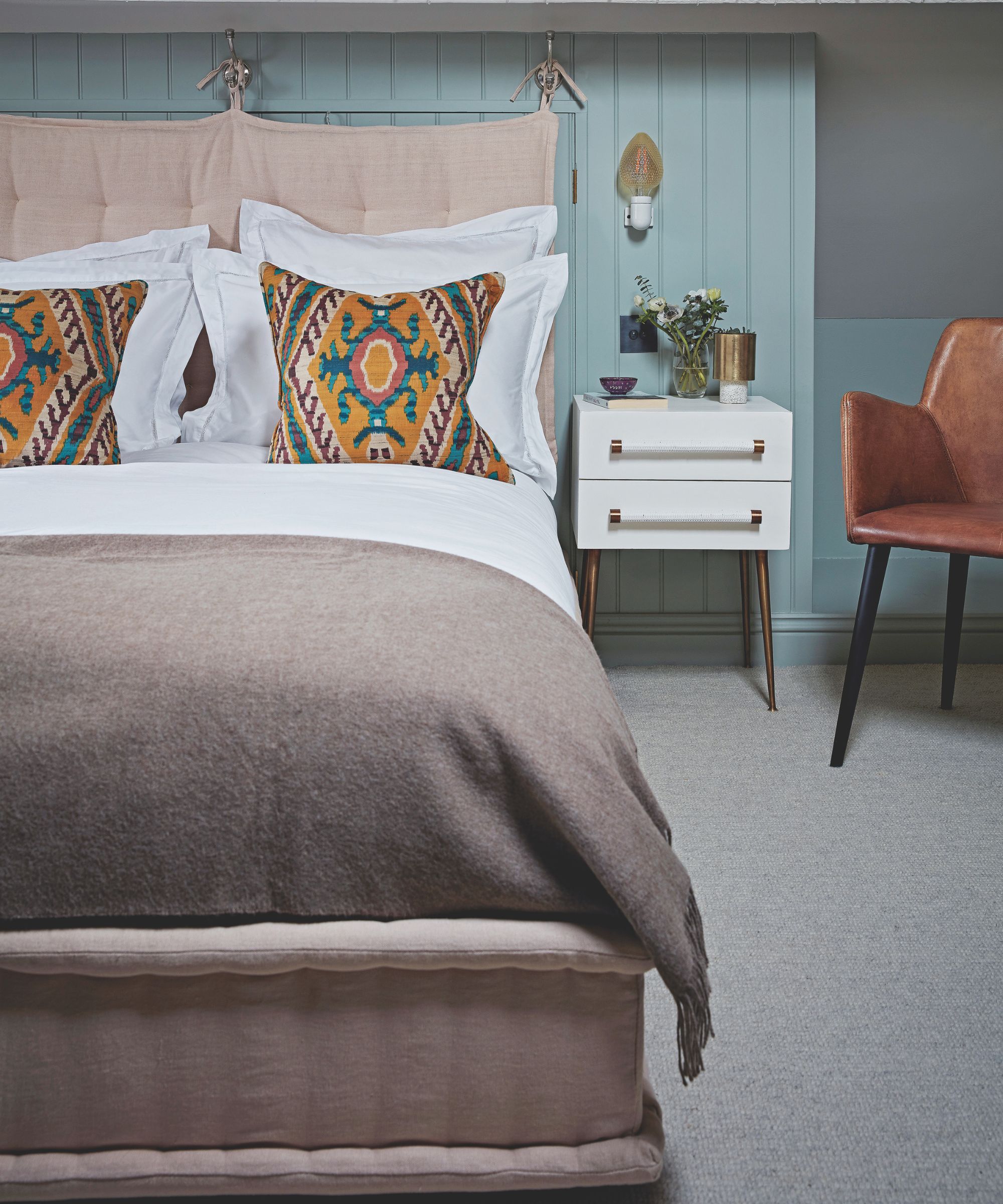 A blue bedroom with panelling behind the double bed, which has white bedding, white pillows, colored cushions and a brown blanket at the foot of it. The bed and headboard are blush pink and soft. To the right of the bed is a white bedside table with a houseplant on top, and to the right of that is a brown leather chair.