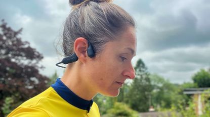 This image shows a woman wearing the Haylou PurFree headphones over her right ear. She has her hair tied up and has a yellow jersey on. There is greenery and a grey sky in the background