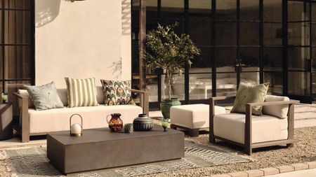 A backyard with brown and white seats with pillows on, a gray table with decor on, a fabric rug underneath, and a white building behind it