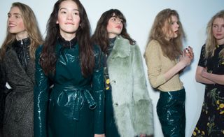 Models waiting in line for a fashion show to start, wearing an elegant gray coat, edgy deep blue leather coat, and pastel green fur coat, from the Topshop Unique A/W 2015 collection.