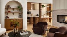 A living room with an arched recessed home bar, an open living and kitchen space with wood accents, a marble floating fireplace, brown armchairs, a marble coffee table, and a white chair 