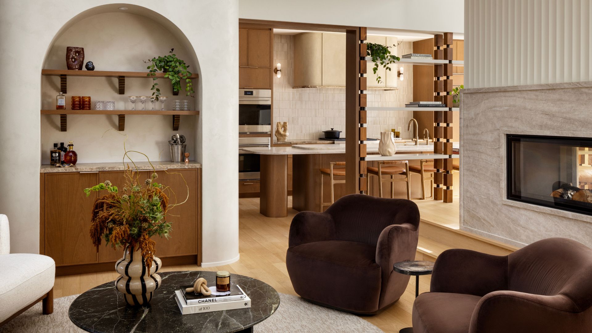 A living room with an arched recessed home bar, an open living and kitchen space with wood accents, a marble floating fireplace, brown armchairs, a marble coffee table, and a white chair 