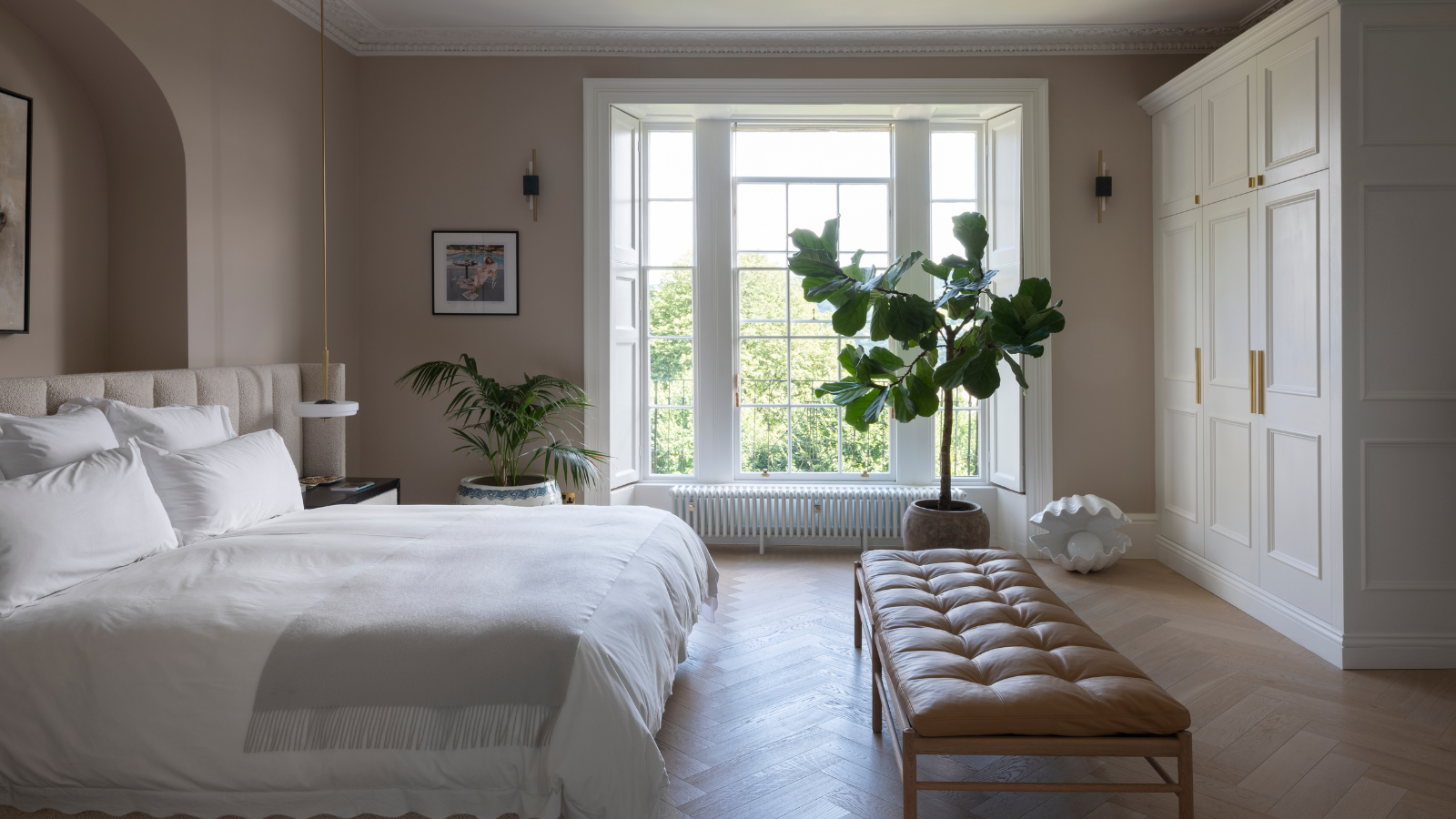 a cozy neutral bedroom with a large bed, a leather bench, a tree in front of the window, and a wall of cabinetry on the right side