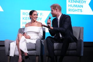 Prince Harry speaks into a microphone as Meghan Markle laughs at what he is saying