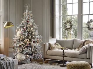 neutral Christmas living room with gold accents - velvet cushions, foot stool, lanterns, flocked faux tree with gold and red decorations, gifts underneath, gold floor lamp, wreath at window