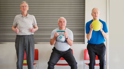 Three men performing chair squats, one is holding a kettlebell, another a yellow ball as they sit back into the chairs