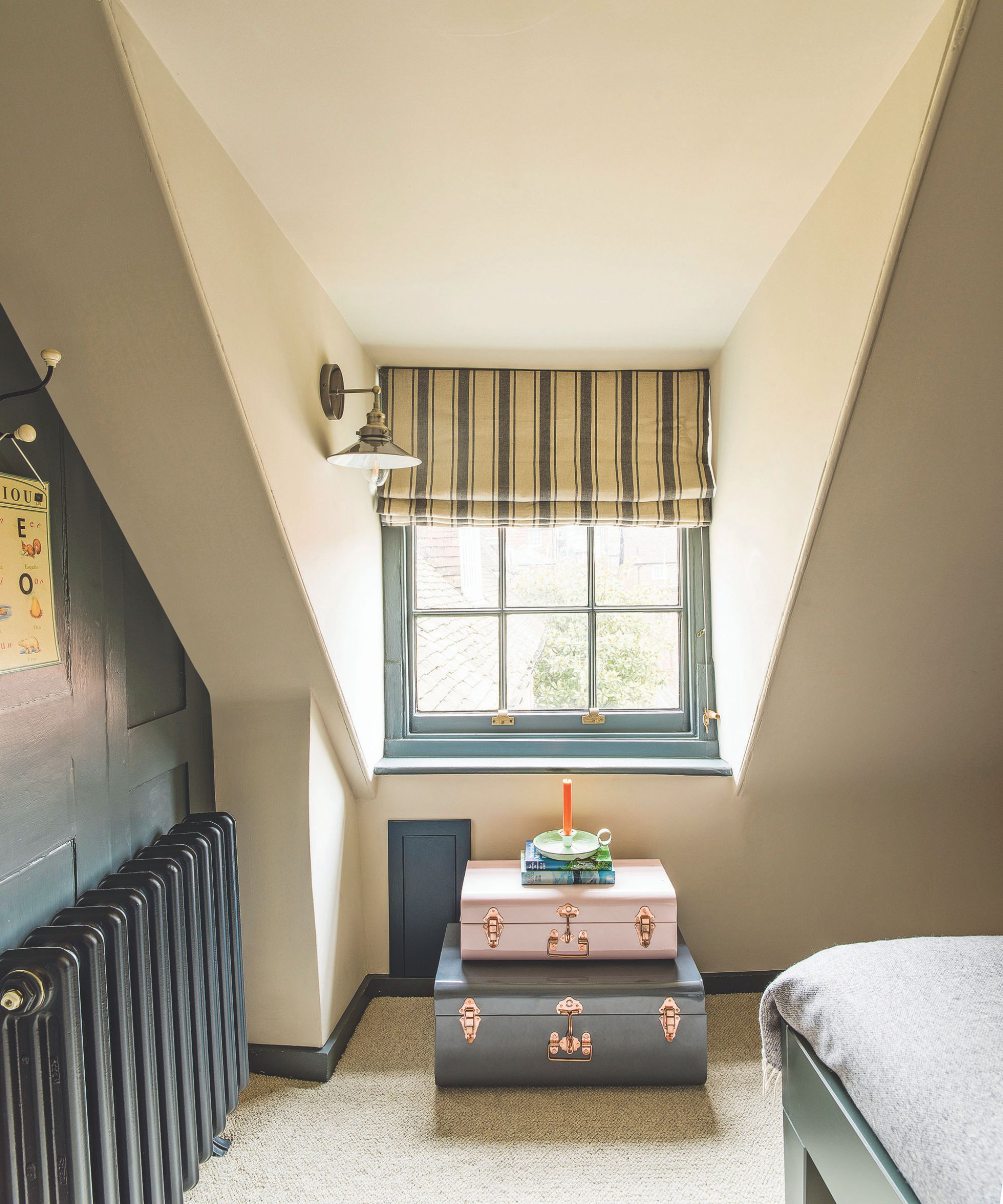 A white attic space with a teal statement wall and radiator, a white and blue striped blind and a stacked suitcase set. To the right is the corner of a blue bedframe with grey bedding.