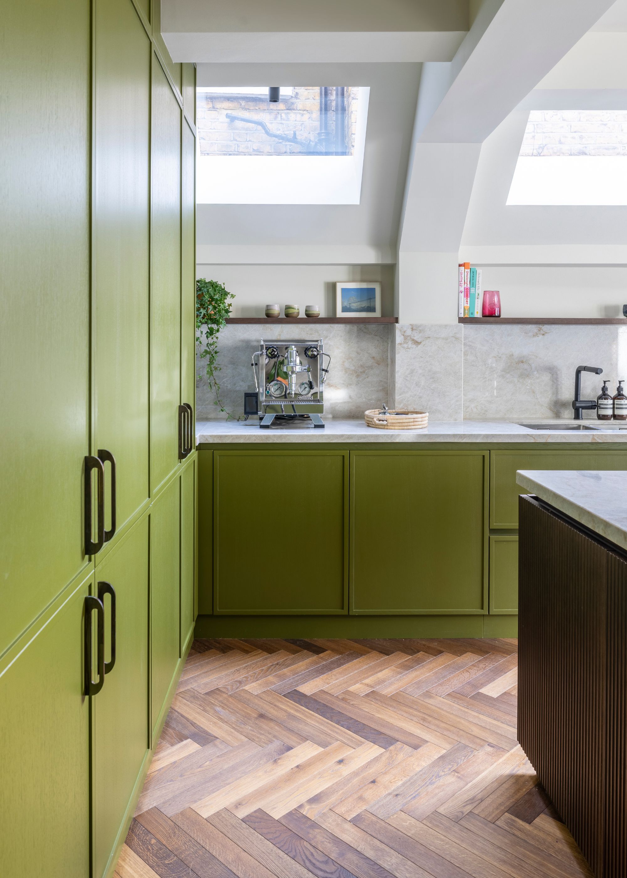 A modern kitchen with warm olive green cabinets, a dark wood kitchen island, white walls with roof windows, and parquet flooring.