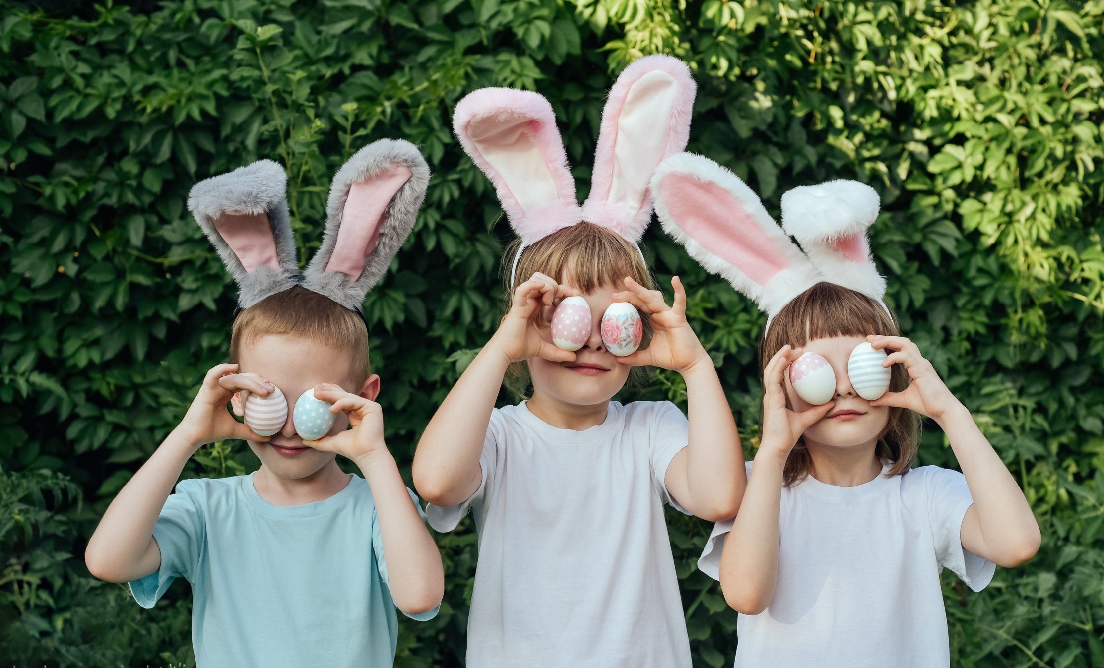 Three kids wearing bunny ears and holding Easter eggs over their eyes for Easter. 