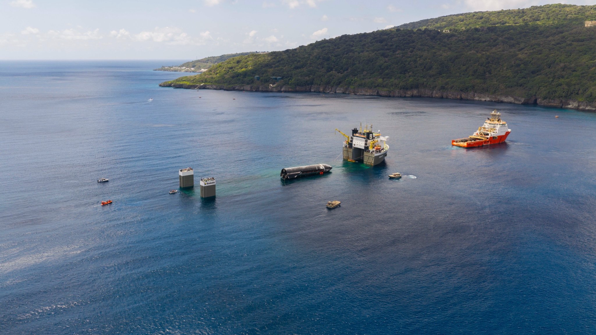 aerial photo of two large ships and several smaller ones in the ocean off a verdant island