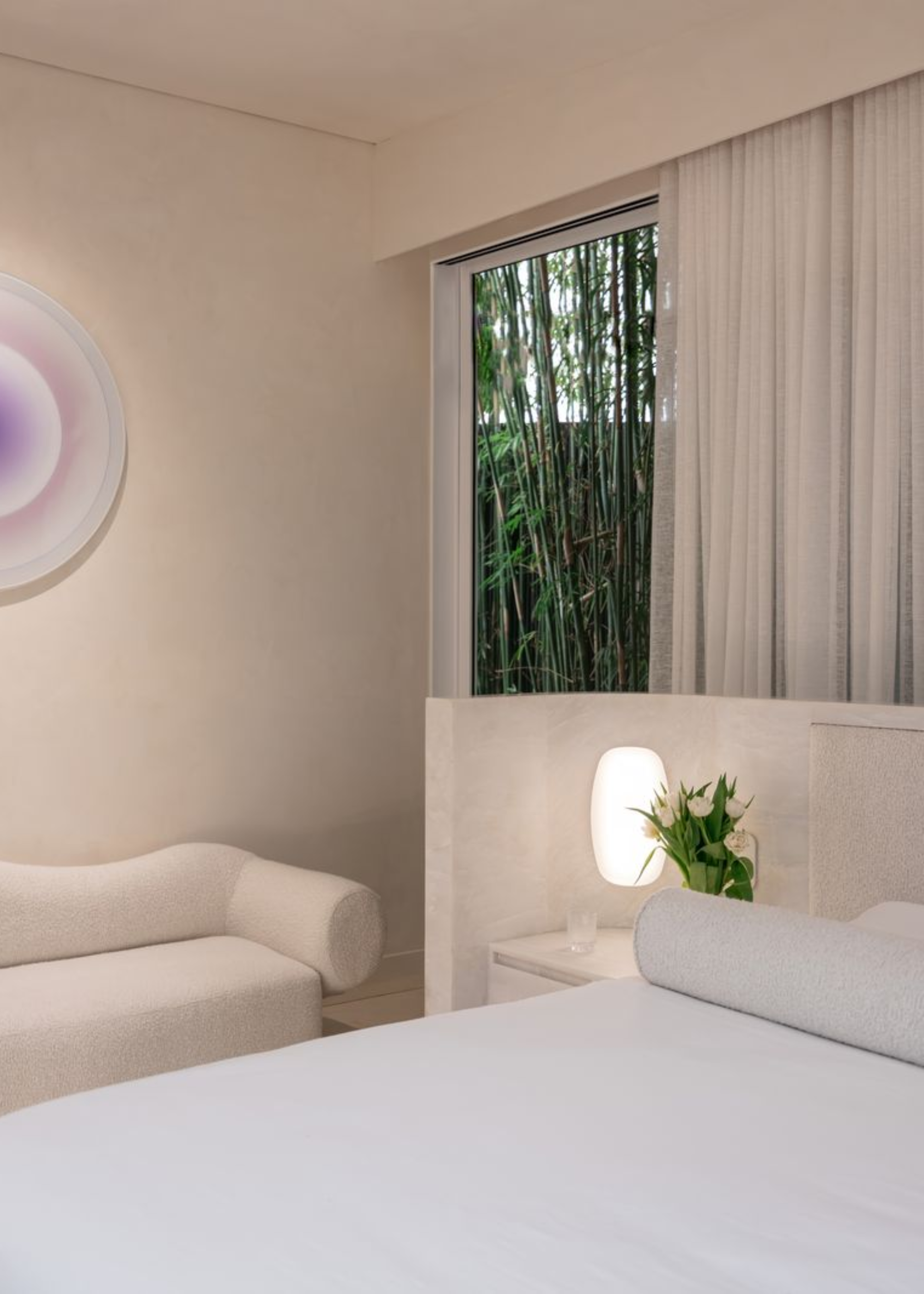 A minimalist white bedroom with sheer window treatments, a purple baclit wall sconce, a white bed and a bedside table with an attached headboard and sconce