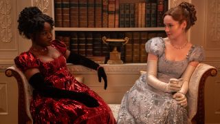 L to R) Masali Baduza as Michaela, Hannah Dodd as Francesca Bridgerton sitting on a couch and looking at each other. 